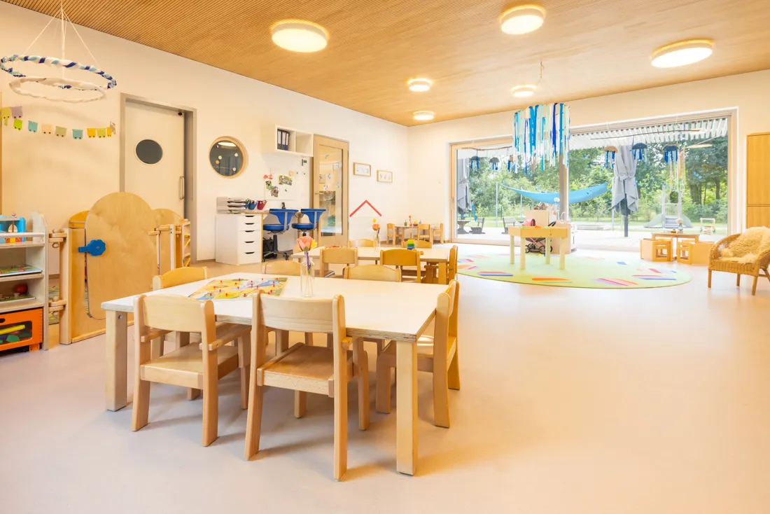 Ein Gruppenraum in einer Kindertagesstätte mit einem Tisch und mehreren Stühlen, bereit für Aktivitäten und Spiele.