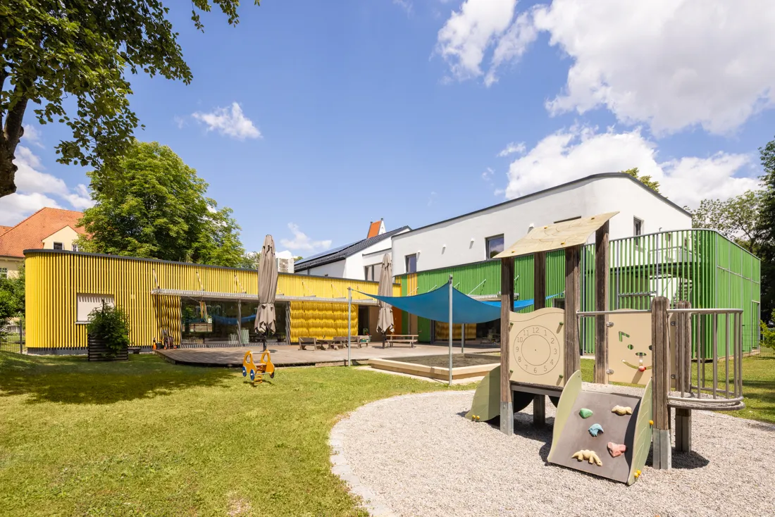 Spielplatz mit einem Baumhaus und einer Rutsche, umgeben von bunten Spielgeräten und der Kindertageseinrichtung St. Mauritius im Hintergrund