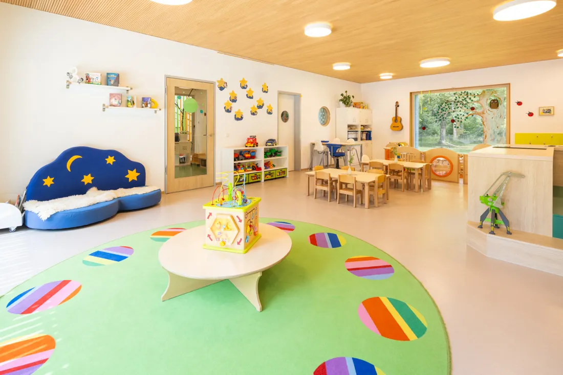 Bunter Gruppenraum des Kindergartens mit buntem Teppich, vielen Spielzeugen und Tischen und Stühlen, ideal zum Spielen und Lernen