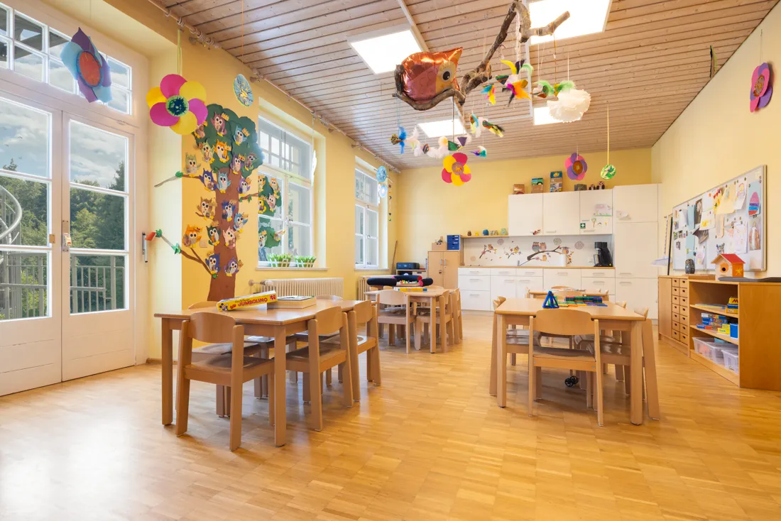 Bunter Gruppenraum mit vielen Basteleien an Wänden und Decken und vielen Tischen und Stüheln zum Spielen, Malen und Lernen