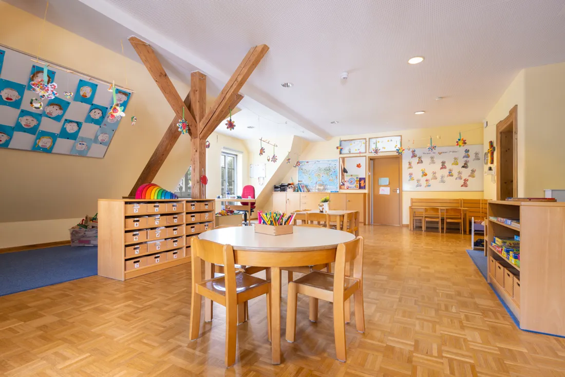 Großer Gruppenraum mit Dachschräge und vielen Tischen zum Malen, Spielbereich auf dem Boden und vielen Regalen voller Spiele und Bastelsachen