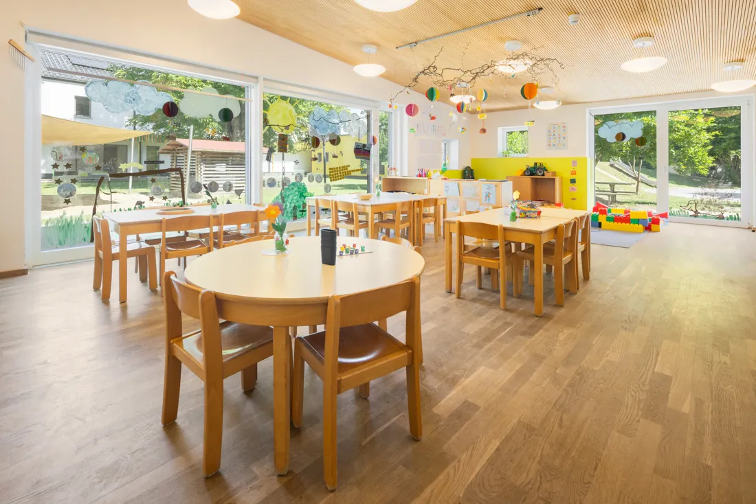 Großer offener Gruppenraum mit großen Fenstern und vielen Tischen und Stühlen zum Malen und Spielen